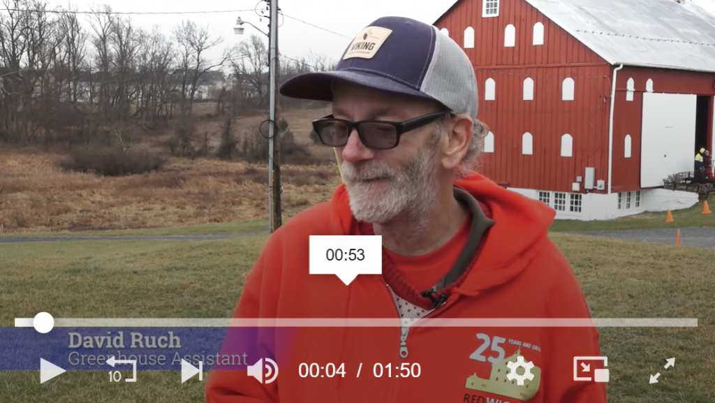 male employee with a blue trucker hat stands in front of red barn, being interviewed about his work on the farm