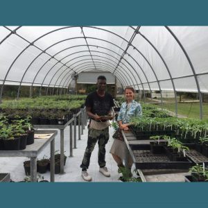 Photo of two people in a greenhouse smiling for the camera. One person holds a clipboard and pencil.