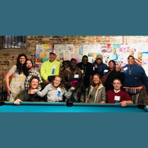 Photo of two employees sitting behind the GROW HUB table at an event, with a customer speaking to them from the other side of the table.
Photo of 13 people posing together in front of a wall covered in art projects.